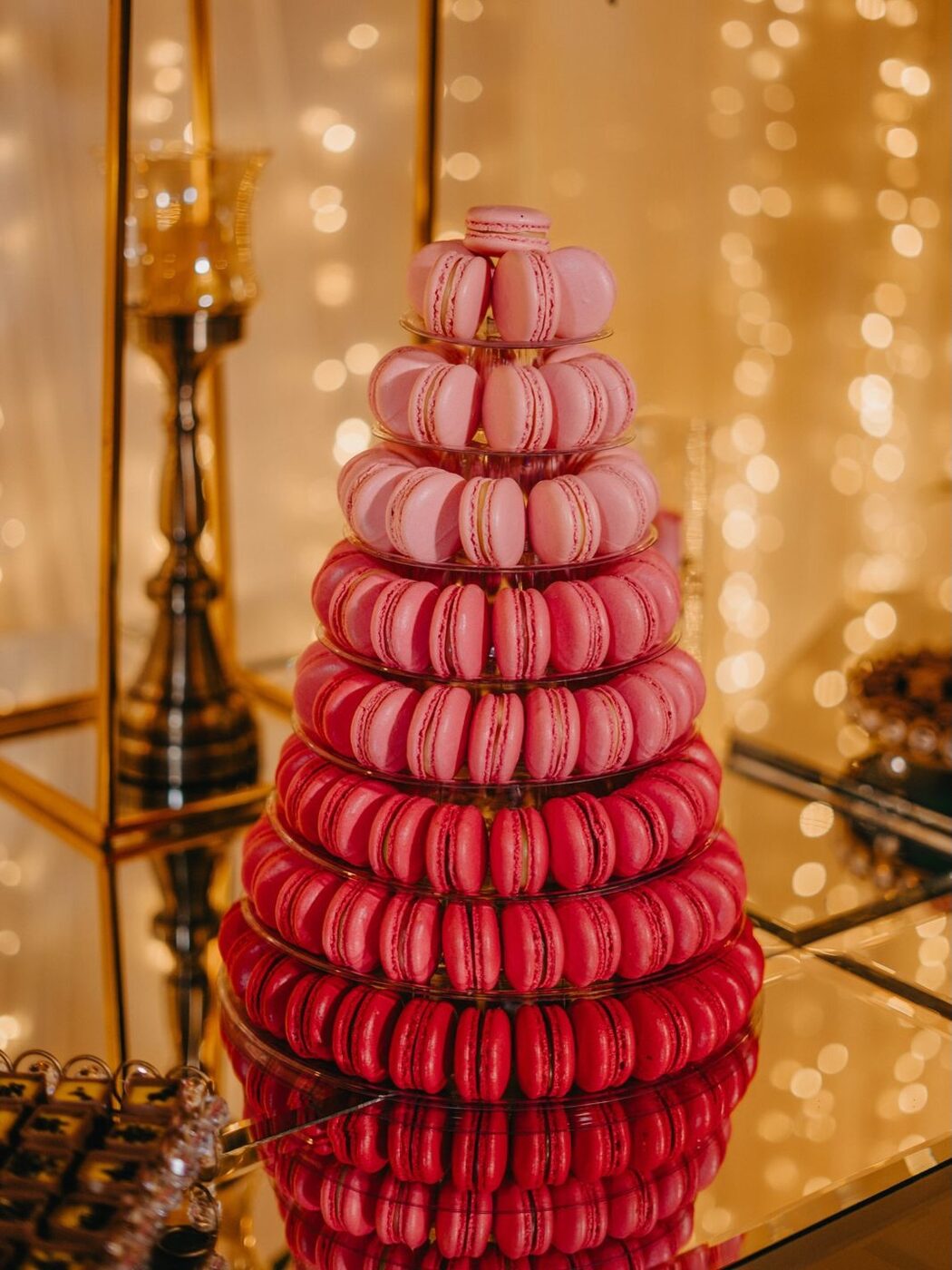 Torre de 10 andares de macarons em degradê de rosa a framboesa, sobre espelho com luzes douradas