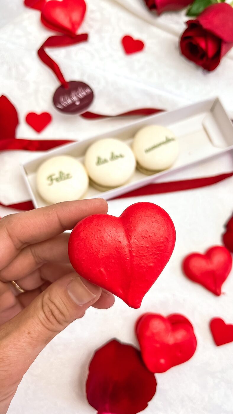 Macaron em formato de coração vermelho segurado pela mão, com caixa de macarons ao fundo